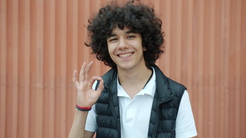 Slow motion portrait of cheerful Middle Eastern teenager showing ok hand gesture and smiling outdoors against wall background. People and emotions concept.