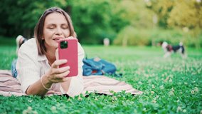 Beautiful adult woman taking a video call on her smartphone while lying on plaid at the park.