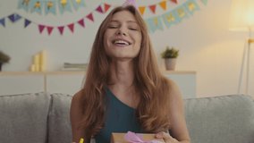 Happy birthday greeting. Close up portrait of excited young lady wearing holiday cap blowing up whistle and offering gift box to camera, dancing at home, slow motion