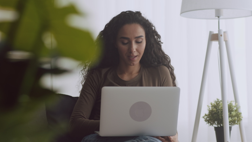 Online win. Youn curly woman web surfing on laptop, entering information and winning, laughing and enjoying victory, sitting in armchair at home, tracking shot, slow motion