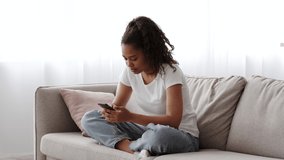 Smiling african american teen girl using smartphone while relaxing on couch at home, black teenage female surfing internet, chatting in social media or texting with friends, slow motion footage