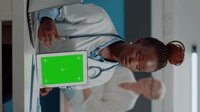 Vertical video: Medic showing green screen display on tablet to senior man sitting at desk in healthcare office. Doctor vertically holding isolated template with mockup background and chroma key