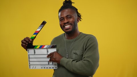 African American second assistant camera clicking clapperboard at yellow background. Portrait of smiling cheerful handsome man posing looking at camera. Filmmaking and video production