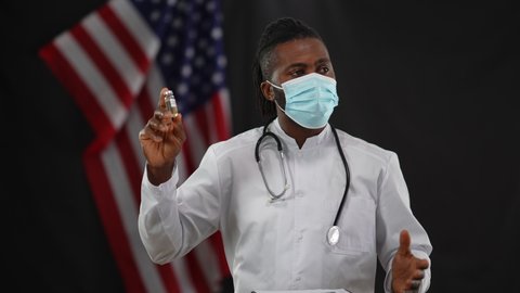 African American male scientist in coronavirus face mask presenting Covid-19 vaccine at press conference in camera flashes. Confident doctor talking and gesturing advertising drugs