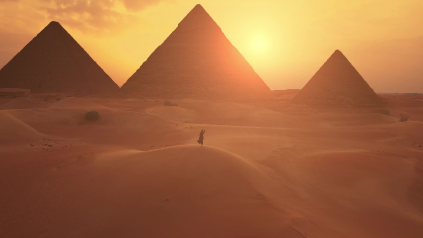 Female traveller looks to the silhouette of pyramids at the sunset in the desert. Aerial shot. Royalty-Free Stock Footage #1078012886