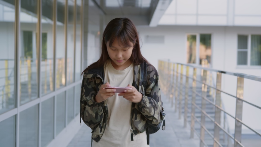 Slider shot of asian teen girl using smartphone walking outside school. Portrait of korean teenage student with mobile phone walking near school building