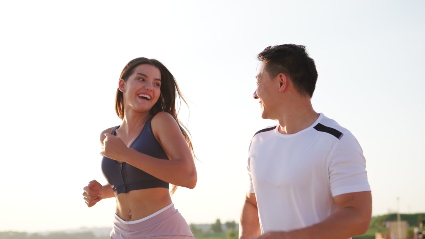 Joyful energetic young sports couple actively training outside, running along the beach and smiling happy to each other. Healthy lifestyle. People and sport.