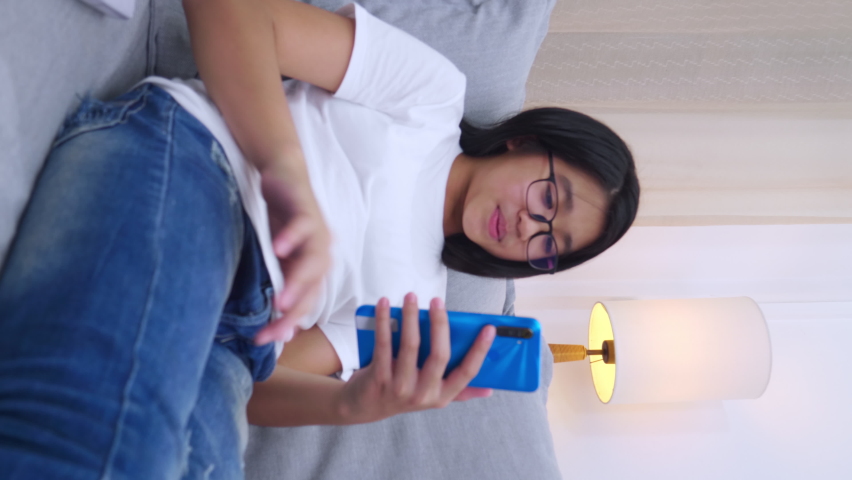 Vertical footage. Happy Asian girl sitting on sofa in living room speak with parent on mobile phone, smiling cute girl use video call by smartphone speak with family. via internet WiFi.