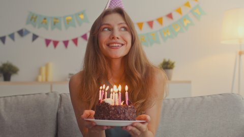 Happy excited woman making cherished wish and blowing candles on holiday cake, smiling to camera, celebrating birthday at home, slow motion