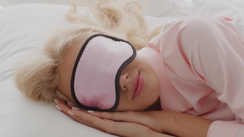 Beautiful young woman wearing sleep mask waking up in bed in the morning, blonde lady taking off mask, stretching arms and opening eyes, awaking in good mood after healthy sleep, slow motion footage