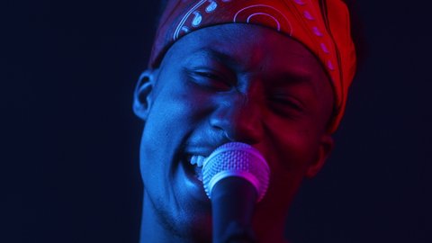 Portrait Of African American Singer Man Singing Song Standing Near Microphone Looking At Camera Posing In Dark Studio Illuminated With Blue And Red Neon Lights. Talents And Professions Concept