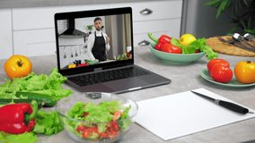 Laptop computer with man food blogger shows slice cucumber tells teaches housewife student stands on kitchen table near vegetables and cutting board with knife, online video call cooking lesson course