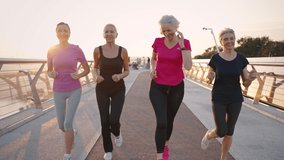 Morning activity for seniors. Group of positive mature women wearing sport clothes running together outdoors, training early on sport ground, slow motion