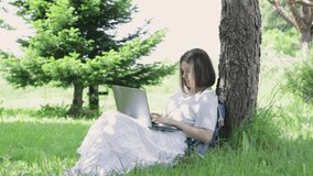 Nice teenager girl sitting in garden nearby tree. Young woman using laptop for tuping on bright sunny summer day. Concept of online learning, distance education and working from home