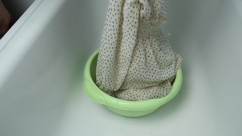 Woman is washing dress from delicate fabric in basin in soapy water with washing powder, laundry by hands at home bathroom. Washing at home, housekeeping and housework.