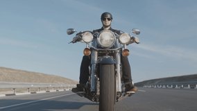 Confident biker in black leather jacket, helmet and sun glasses rides along the highway on cool motorcycle, front down view