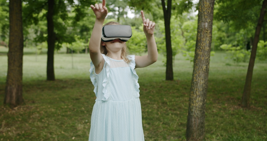 Happy Girl play with modern virtual reality glasses in park in blue dress