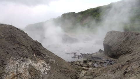 Water boils in a hot spring. Bubbles and splashes above the surface. Steam and haze fly into the sky. Green vegetation on the hillside. In the foreground there are picturesque boulders. Kamchatka