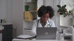 Doctor telemedicine patients service. Spbas Pretty black lady therapist in uniform with stethoscope closes modern laptop sitting at table in clinic