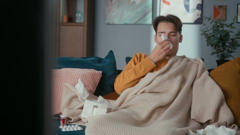 Young man having cold is sitting on sofa, watching TV, sneezing into his handkerchief, taking pills. Being lockdown, on sick leave, coronavirus, self-isolation, home treatment.