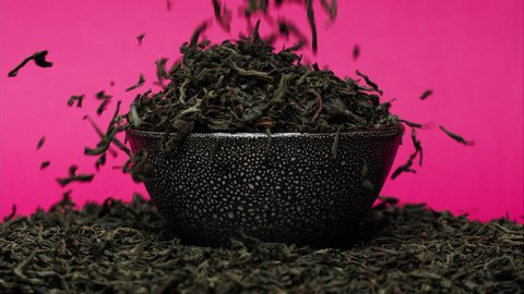 Close-up of falling down dried tea leaves into black bowl on pink background. Shooting of ceylon tea in studio. Macro footage of pouring out brewed tea into pile. Food and drink cooking video. 