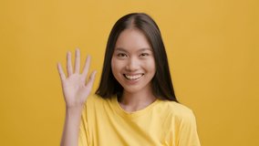 Hello. Cheerful Asian Millennial Woman Waving Hand Playfully Smiling To Camera Posing Over Yellow Studio Background. Headshot Of Happy Chinese Female Gesturing Hi Greeting Concept