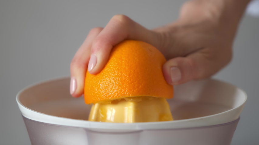 Woman is squeezing oranges using electric juicer in the kitchen, hand closeup. Healthy vegan ripe fruit juice, natural vitamins from food. Making orange juice pressing half of orange.