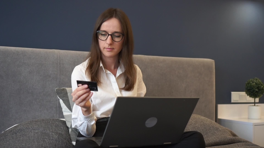 Pretty girl is making online payment holding bank card using modern laptop at home sitting on bed.
