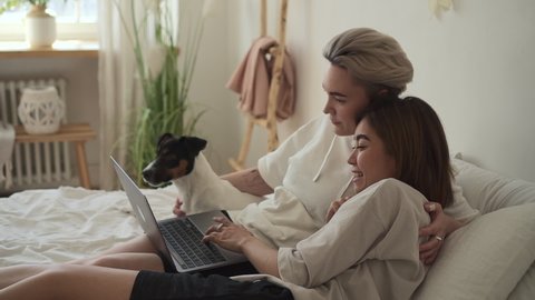 Lesbian girfriend use laptop while lying on bed surfing online, discuss internet together Spbd. Same sex lgbt female couple relax with cute dog. Concept woman, love, home