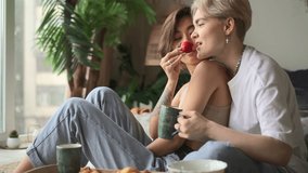 Lesbian couple eats and has fun. Spbd Happy woman and Asian girlfriend with delicious strawberry and cup sit on bed in hotel room in lazy morning - Powered by Shutterstock - Get 15% off with code: PIKWIZARD15