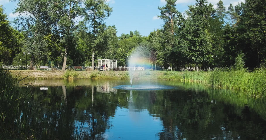 working fountain in the middle of a city pond, against the background of a family with children, a general plan in a park with green trees, on a clear day, in slow motion