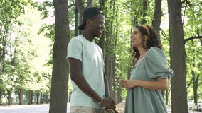 Young Afro-American man and brunette white woman do high-five of consent and laugh against green trees in city park on sunny day