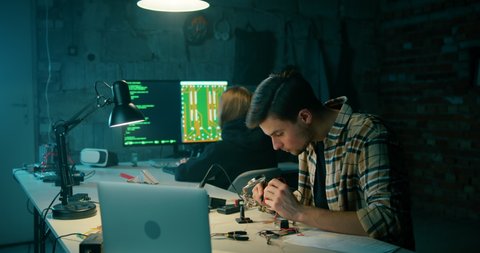 Man Solders in Electronics Workshop. Hacking Crime, Cyber War or IT Startup Concept. Young Programmers Working on Technology Project in Dark Garage or Loft Office. 4K Wide Hand held shot