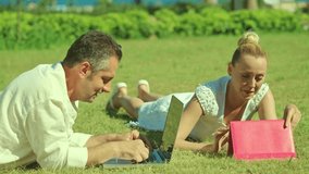 Beautiful woman and her boyfriend working with tablets and computers on green grass. Young woman works with tablet computer while man works on laptop. Remote work concept.