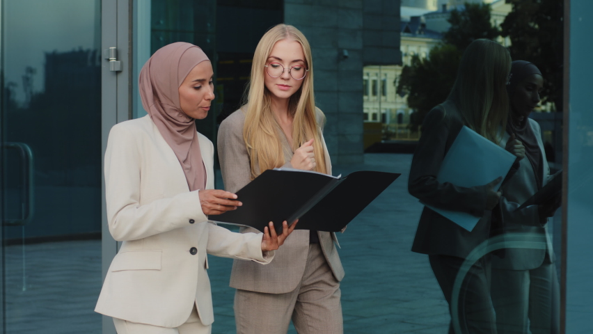 Two diverse businesswomen walking along street holding folder with documents communicate before meeting, discuss report. Indian girl in hijab consults with colleague young European woman during break Royalty-Free Stock Footage #1078098353