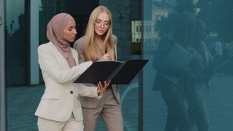 Two diverse businesswomen walking along street holding folder with documents communicate before meeting, discuss report. Indian girl in hijab consults with colleague young European woman during break – Video có sẵn