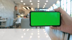 Asian man's hand holding a mobile phone with chroma key green screen display at a coffee shop, video calling, using zoom meeting online app, work from home, working remotely, social distancing