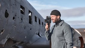 Beautiful steady panning shot, of an attractive male, filming outside the Solheimasandur plane wreck in Iceland using a vintage video camera, with a glimpse of the blue skies in the backdrop.