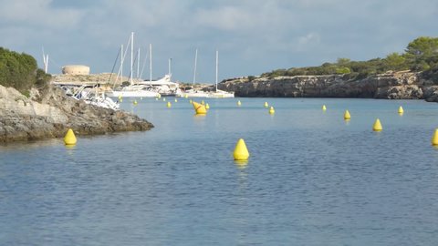 Calm view at sunrise of creek Cala Santandria in Menorca with yellow buoys and boats, blue sea and surrounding rocks.