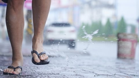 A flip flops feet carefully running on an urban paving footpath during a day, low angle shot on female legs in casual clothing style, a car parked on street road, splashing purity water puddle  