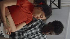 Vertical video: Interracial couple sitting on sofa using laptop in living room. Multi ethnic married people looking at screen together and talking. Mixed race husband and wife at home on sofa with