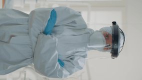 Vertical video: Portrait of caucasian dentist prepared for dental surgery at oral clinic, wearing protection suit with mask, face shield, gloves and coverall. Stomatology specialist looking at camera