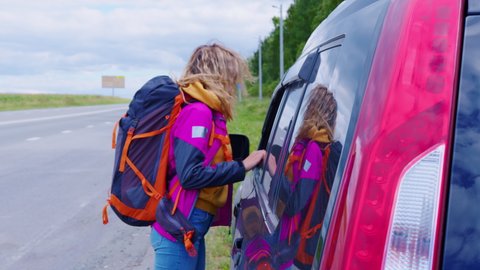 Hiker blond girl with backpack asked for passengers to crossover driver. Hitchhiker woman stopped car on road. Tired tourist lady with knapsack gets into car on roadside. Travel, hitchhiking concept
