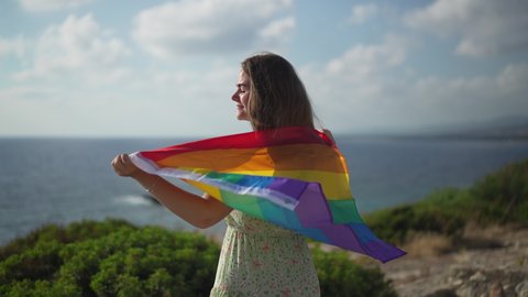 Cheerful Caucasian LGBT woman with rainbow flag smiling stretching hands in slow motion admiring sunset sunrise over Mediterranean sea. Positive happy carefree lesbian enjoying summer vacations