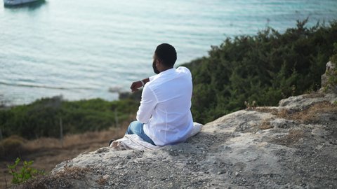 Back view of relaxed African American LGBT man sitting on cliff admiring beauty of blue Mediterranean sea. Happy carefree gay tourist resting on vacations. Tranquility and freedom concept
