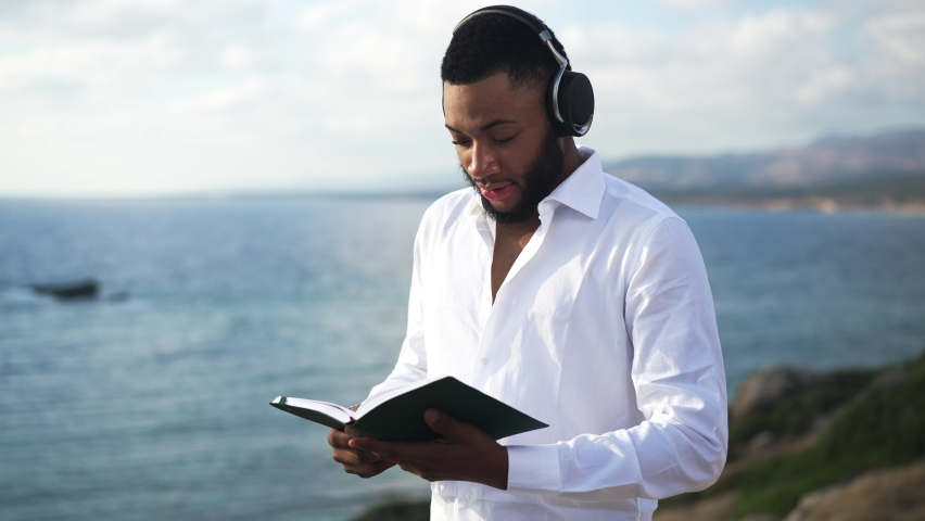 Handsome thoughtful African American man reading book on Mediterranean sea coast at sunrise looking at seascape. Portrait of confident intelligent LGBT person enjoying hobby outdoors