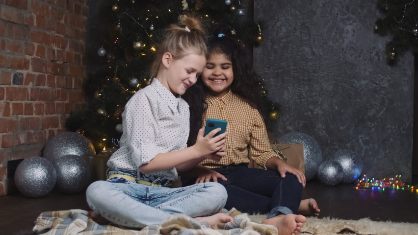 Two pretty young african and caucasian girls sitting near a New Year or Christmas tree and communicate via video link in a smartphone.