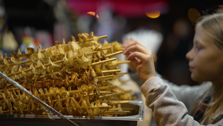 Spiral potato chips on skewers, blurred girl on background choosing one and getting change from seller. Snacks at street fair. Concept of food