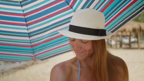 beautiful girl in swimsuit and white hat smears her shoulder with sunscreen while sitting under sun umbrella on beach with sandy beach. Rest by ocean and sea. Protection skin. Vacation, travel