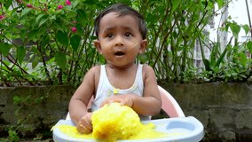 Asian toddler boy is making fun of big yellow ripe mangoes. Baby boy eating ripe mango fruit sitting in the baby tray-chair close-up view. Baby eating fruit in the garden. 4k video.